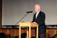 Detailaufnahme eines Mannes am Rednerpult. Er trägt einen schwarzen Anzug und richtet seine Rede an das Publikum.